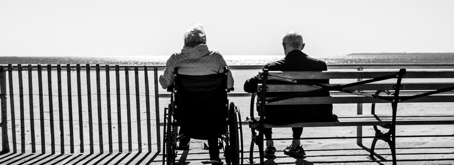 Black and white photo of an elderly couple sitting on a bench and wheelchair looking out at the beach